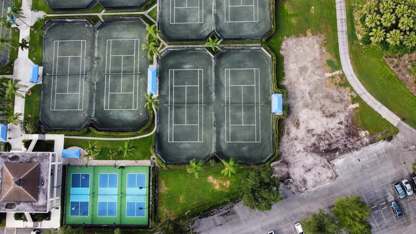 Aerial view of green tennis courts beside a road, with a small blue pool and grassy area nearby