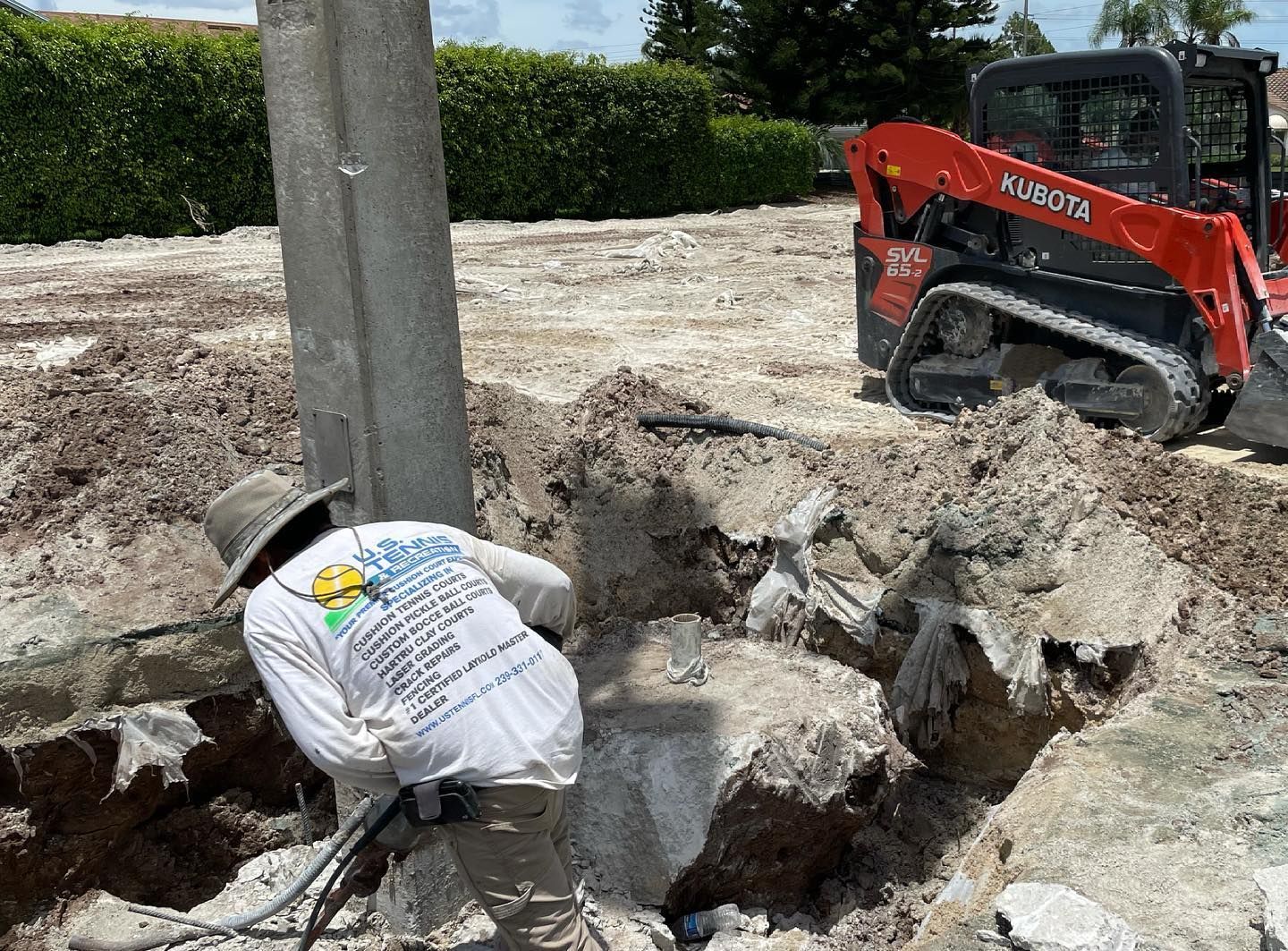 Worker in white shirt breaking concrete beside a pole, with a red Bobcat skid steer in the background.