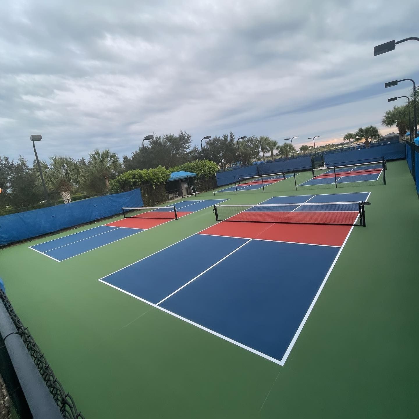 Outdoor pickleball courts with blue and red playing surfaces on a green court under a cloudy sky