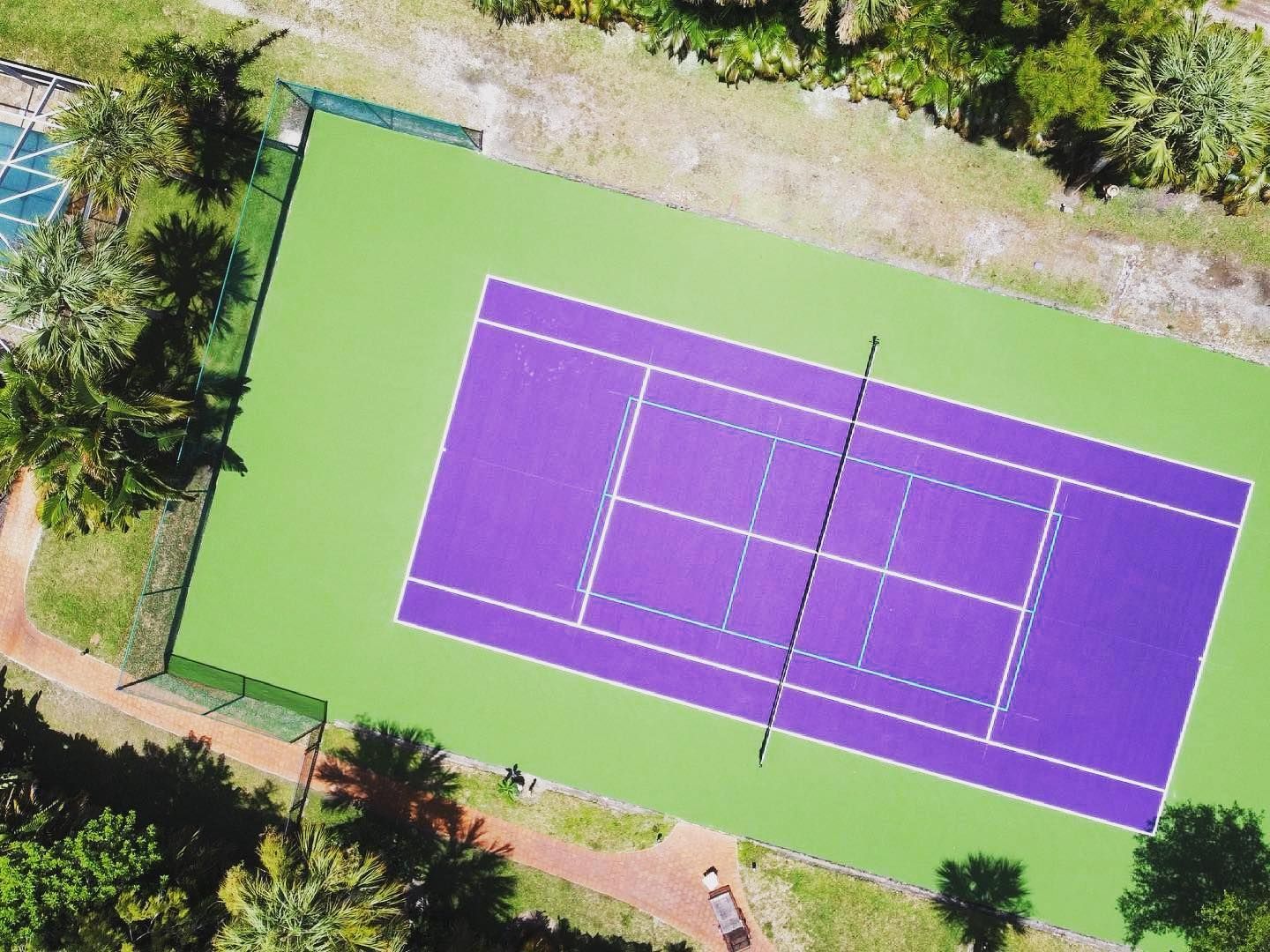 Aerial view of a purple tennis court with green outer area, surrounded by trees and paths.