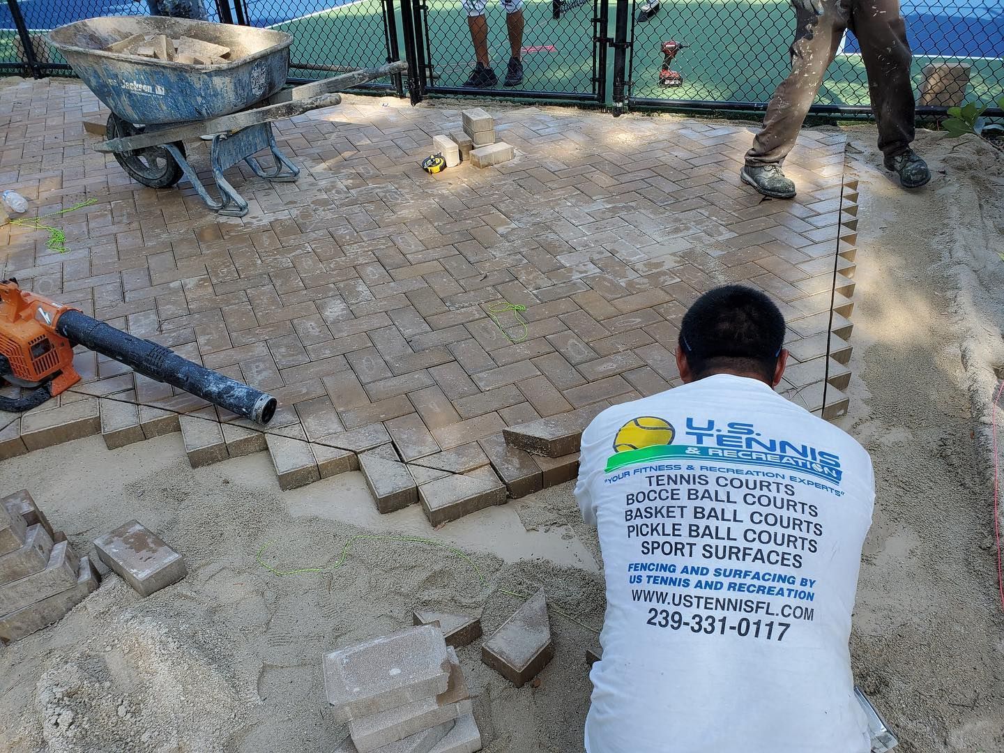 Worker crouches on brick walkway, using a blue hose amid scattered pavers and tools.