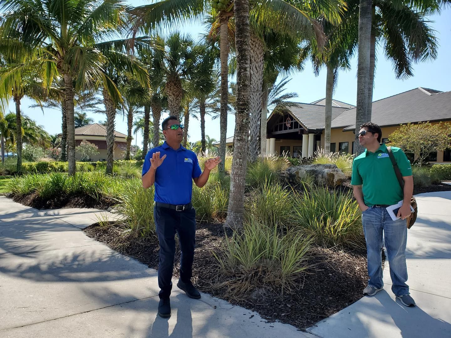 Two people standing on a sunny walkway beside palm trees and a building with a metal roof.