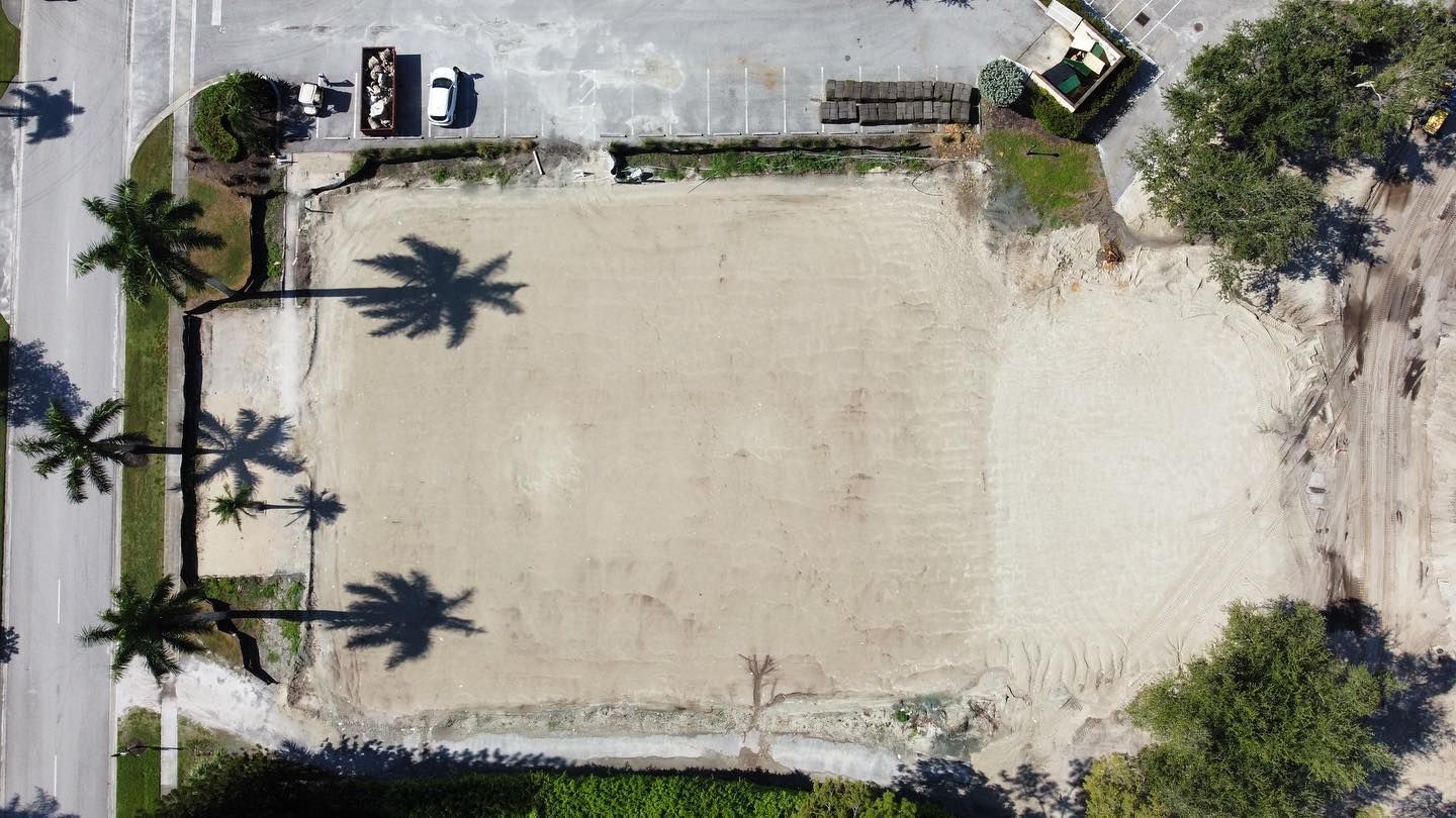 Aerial view of a sandy rectangular court bordered by palm trees and parked vehicles along the top edge