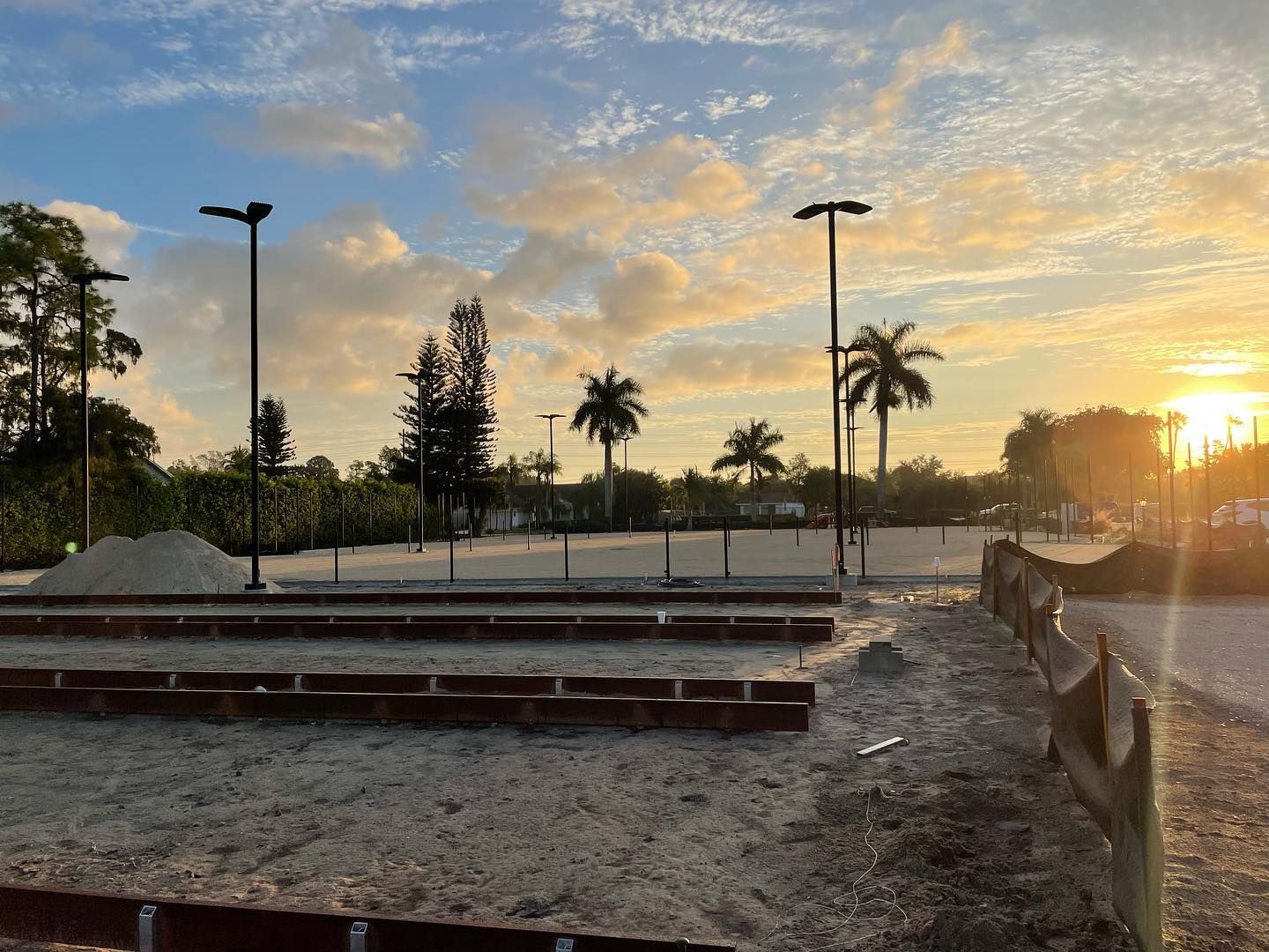 Sunset over an outdoor construction site with benches, light poles, and palm trees.