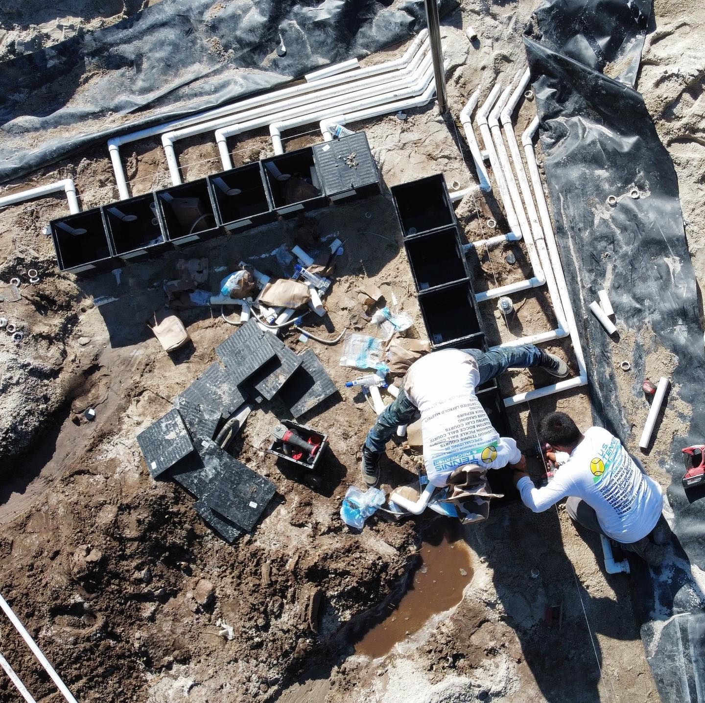 Workers digging in a rocky excavation beside black pipe segments and white drainage tubing.