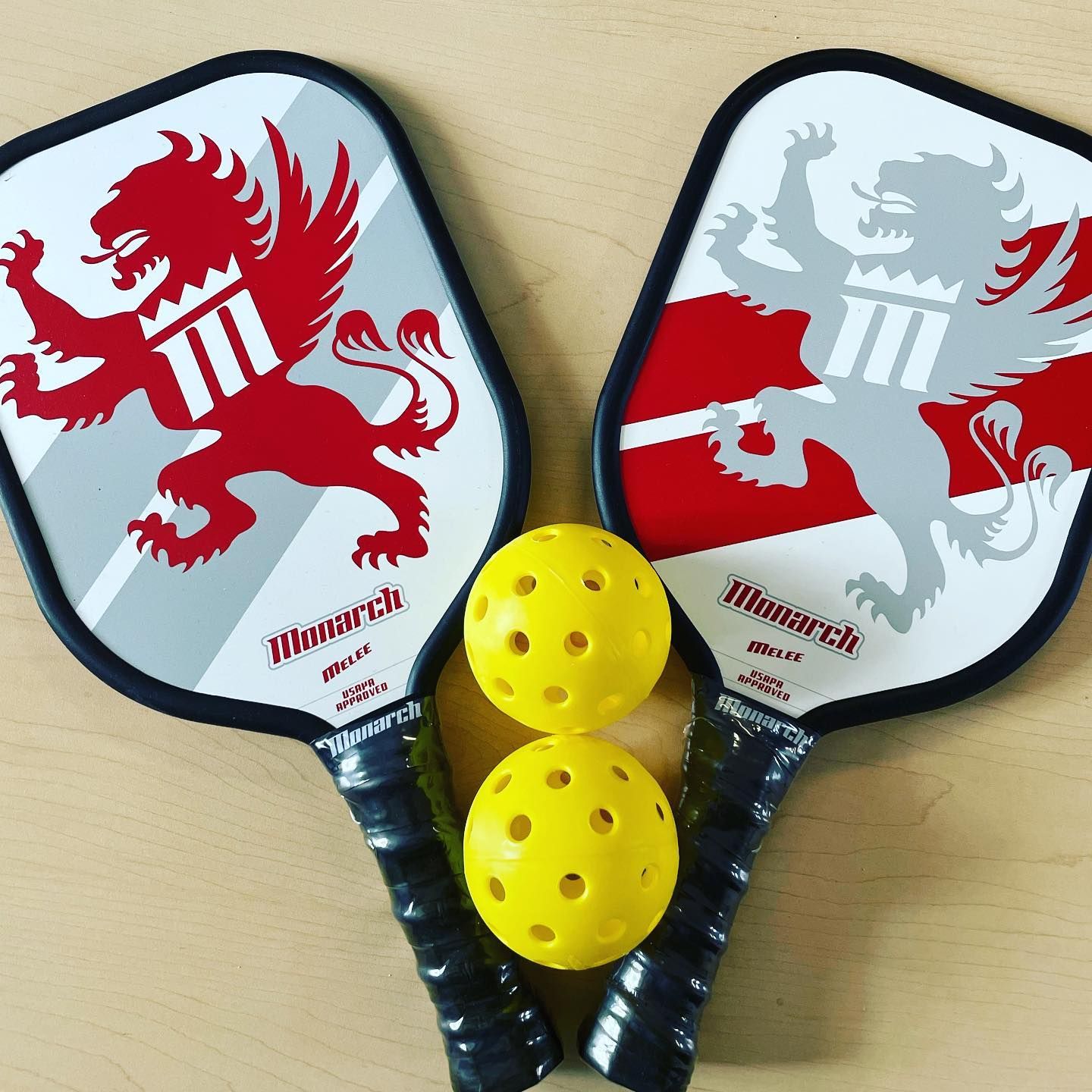Two black padel rackets with red dragon designs and two yellow perforated balls on a light wood table