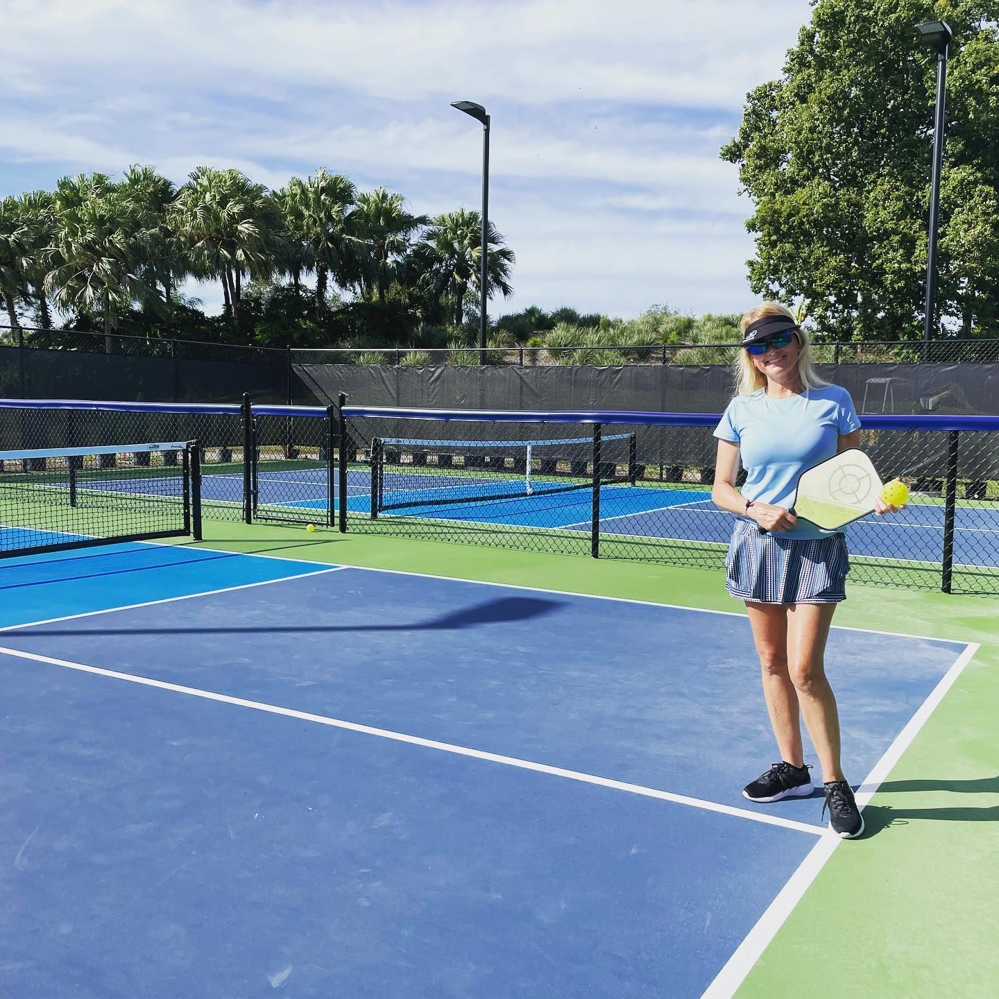 Tennis player holding a racket on a blue court beside a green sideline, with netted courts and trees behind.