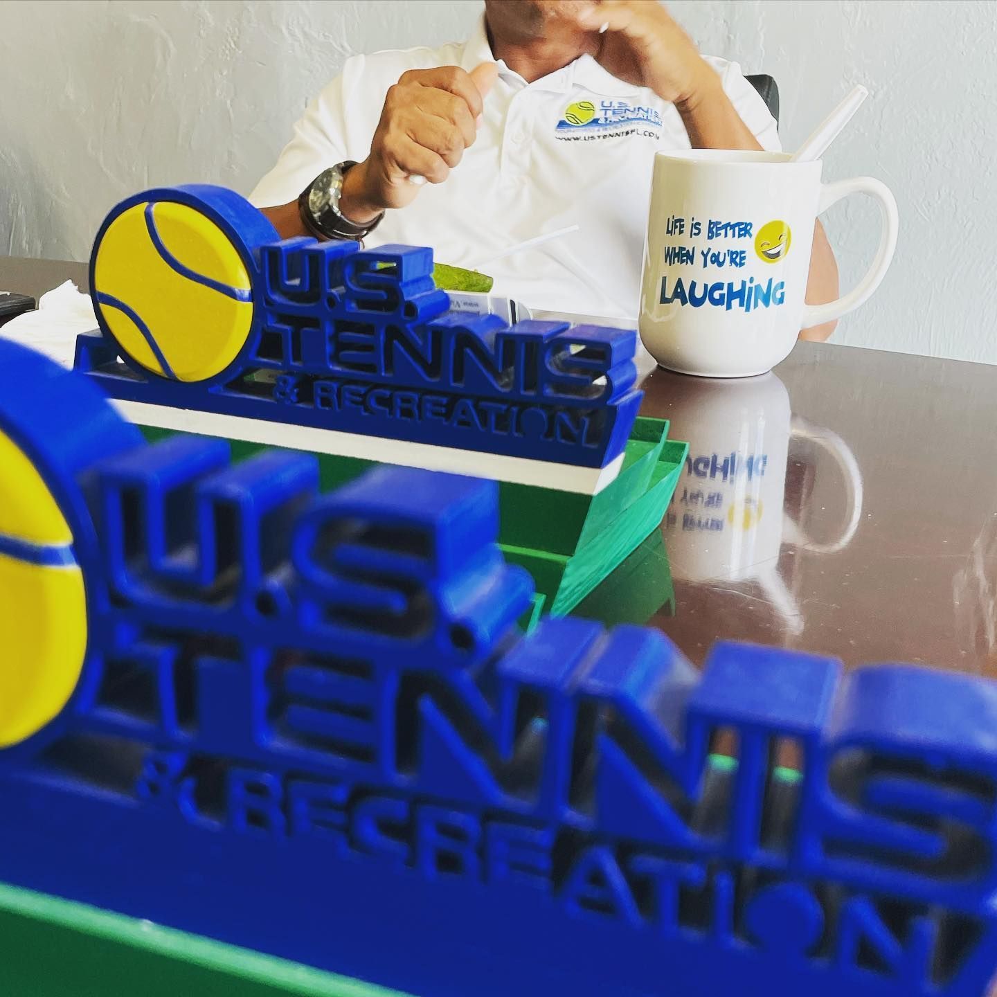 Blue 3D tennis sign beside mugs with a man holding a tennis ball in the background