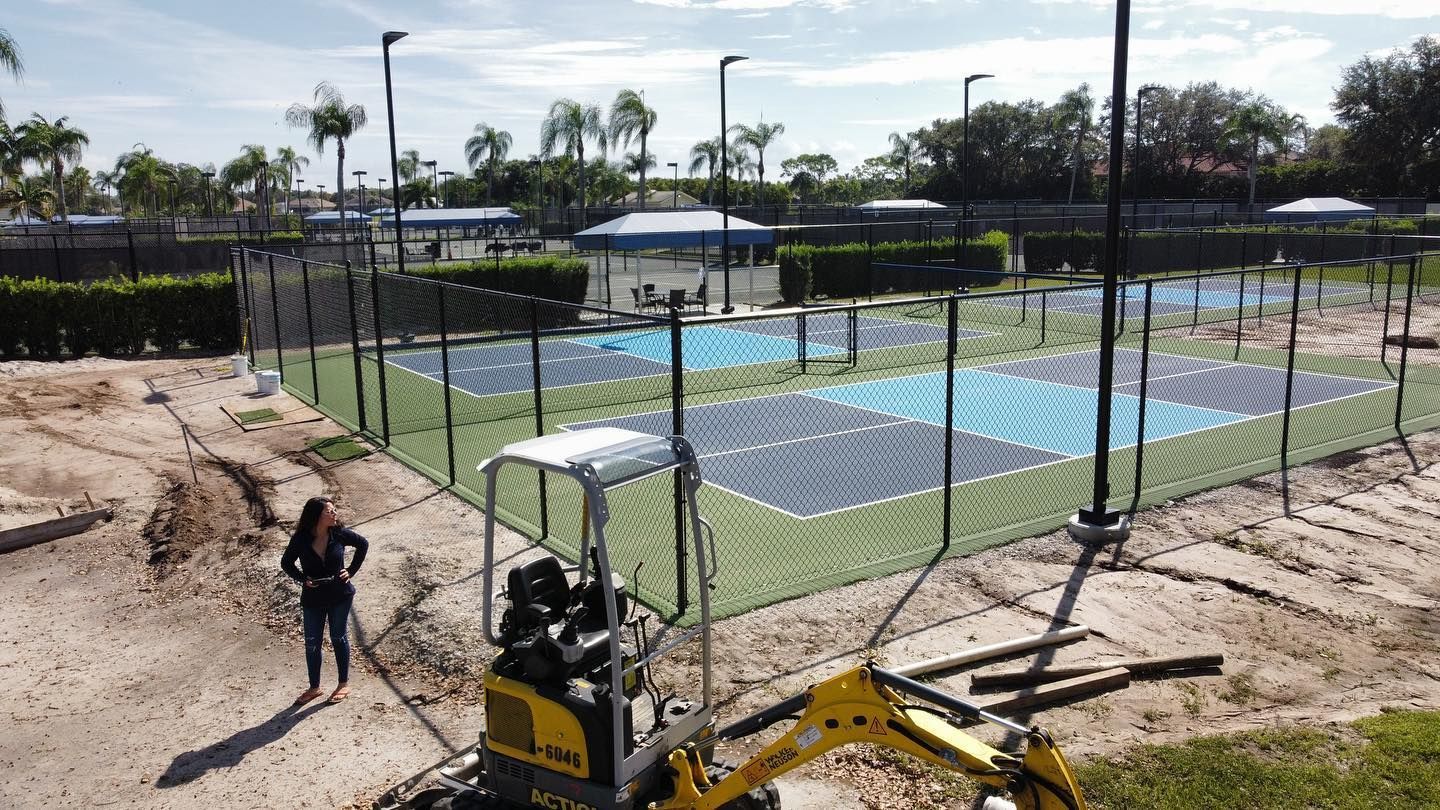 Tennis courts behind chain-link fence with a construction worker and mini excavator in the foreground