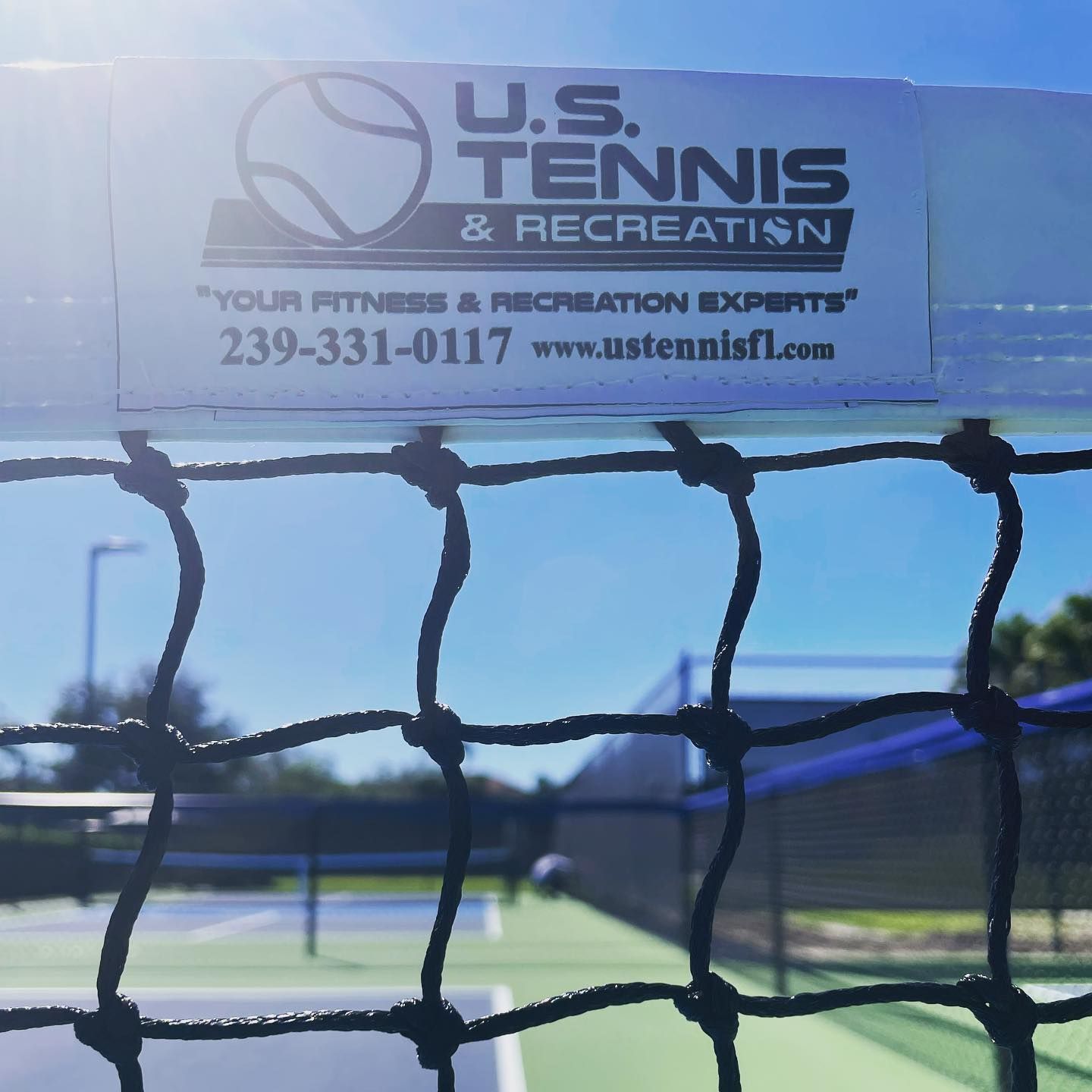 Tennis net with a U.S. Tennis & Recreation banner against a bright blue sky and court backdrop