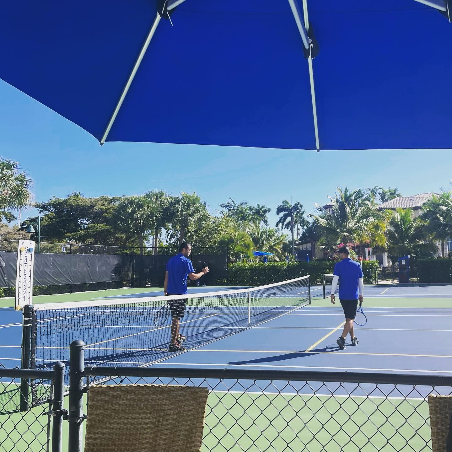 Two people in blue shirts playing tennis on an outdoor court under a large blue umbrella.