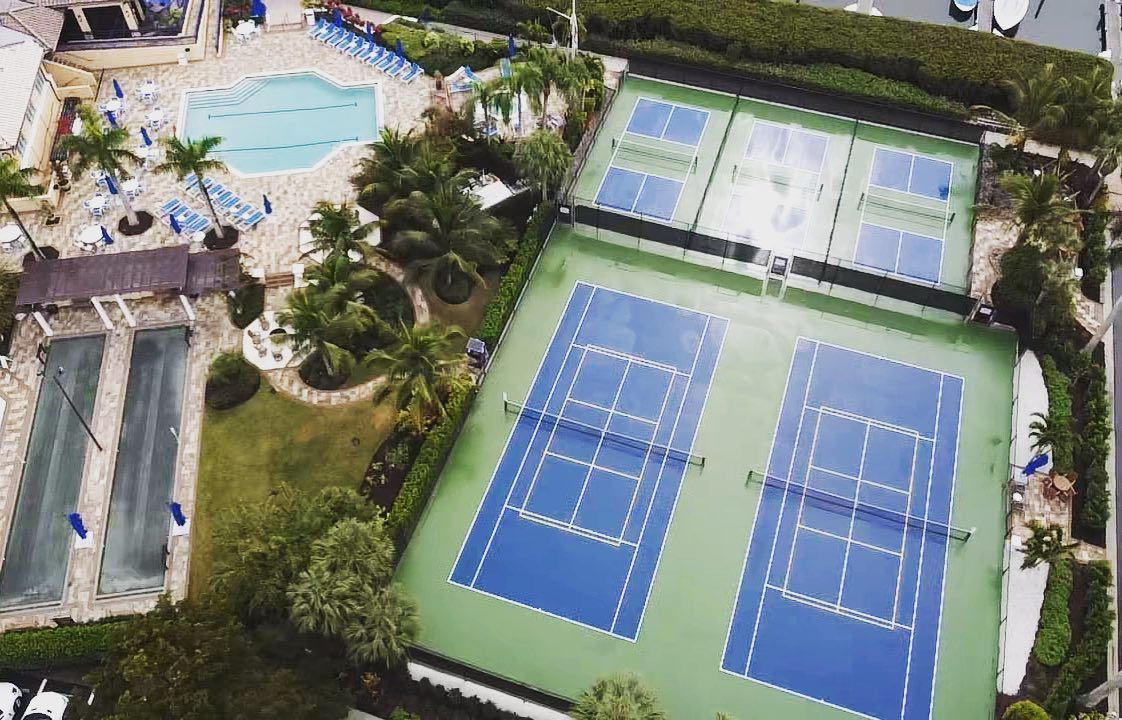 Aerial view of a resort with blue tennis courts, a swimming pool, and palm trees.