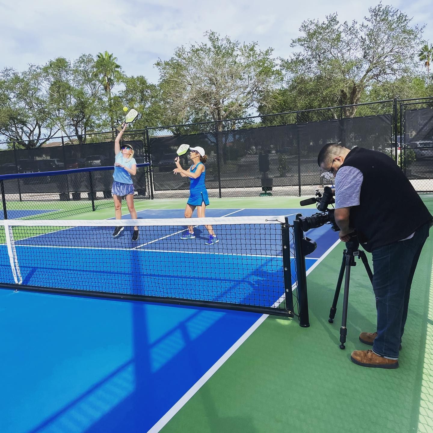 Two people play pickleball on a blue outdoor court while a cameraman films from the sideline.