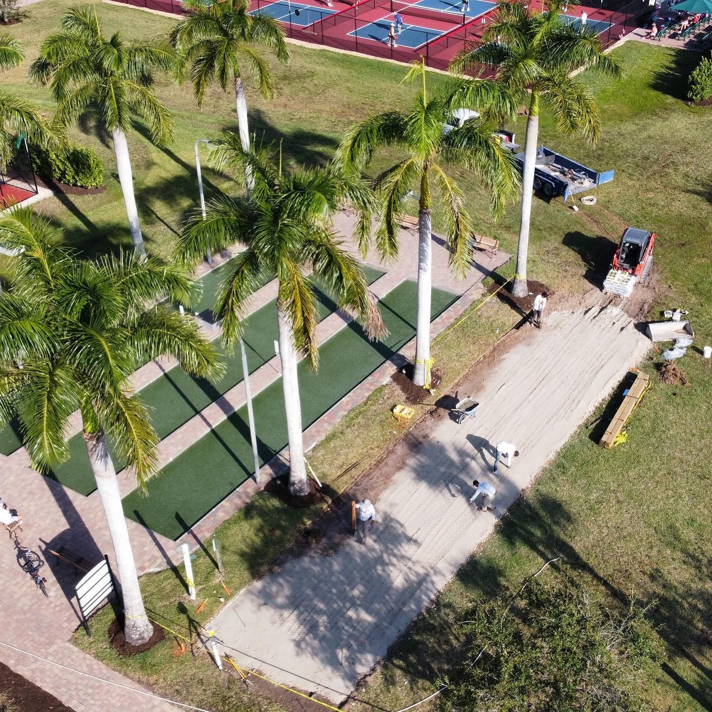 Palm-lined park walkway beside a road, with a green court and red tennis courts in the background