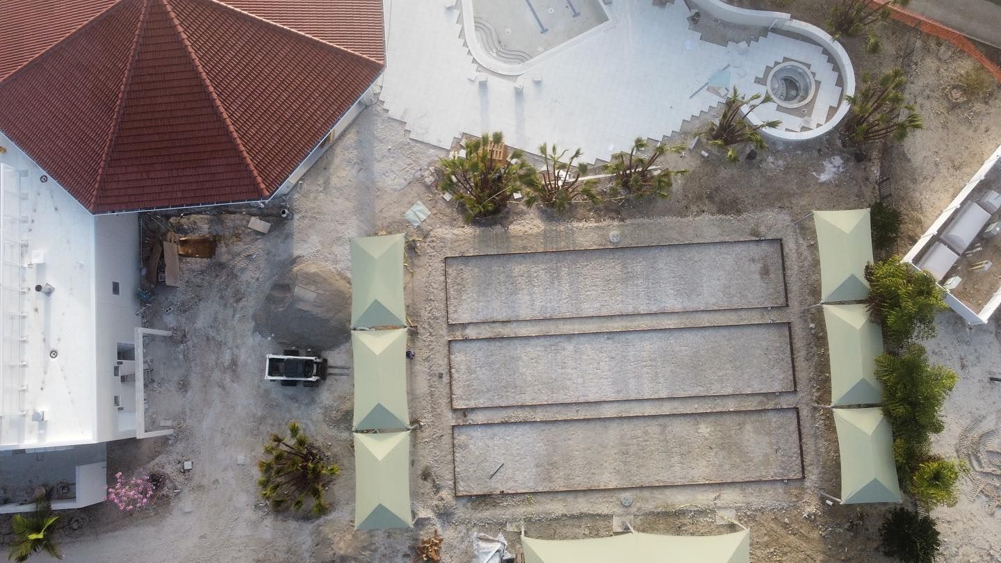Aerial view of a backyard pool area with a small roofed patio, pathways, and a covered rectangular structure.