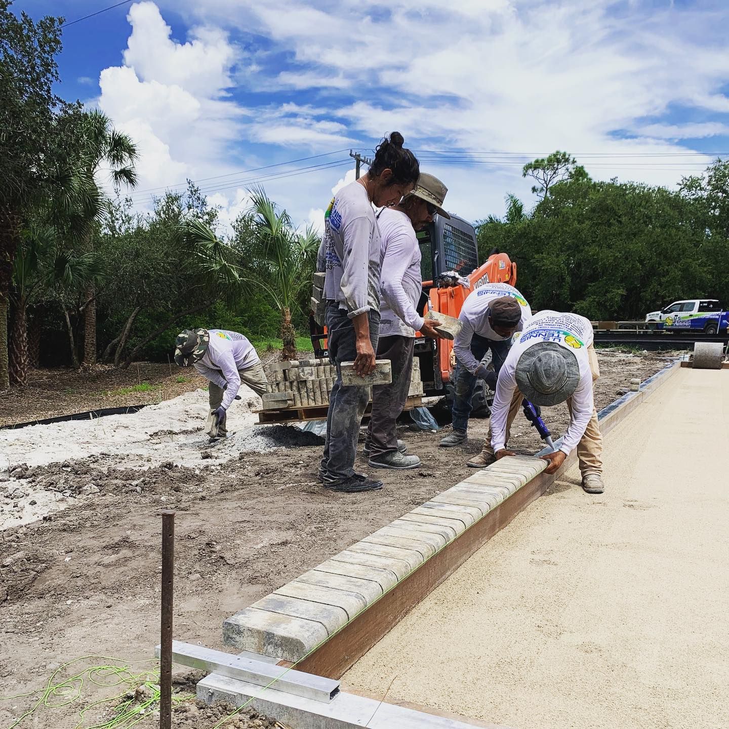 Workers laying concrete curbs beside a newly paved road under a cloudy sky