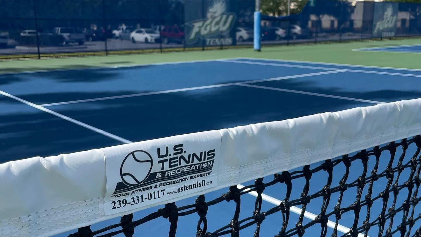 Tennis court viewed from behind the net with a U.S. Tennis sign on the net and blue courtside fencing in back