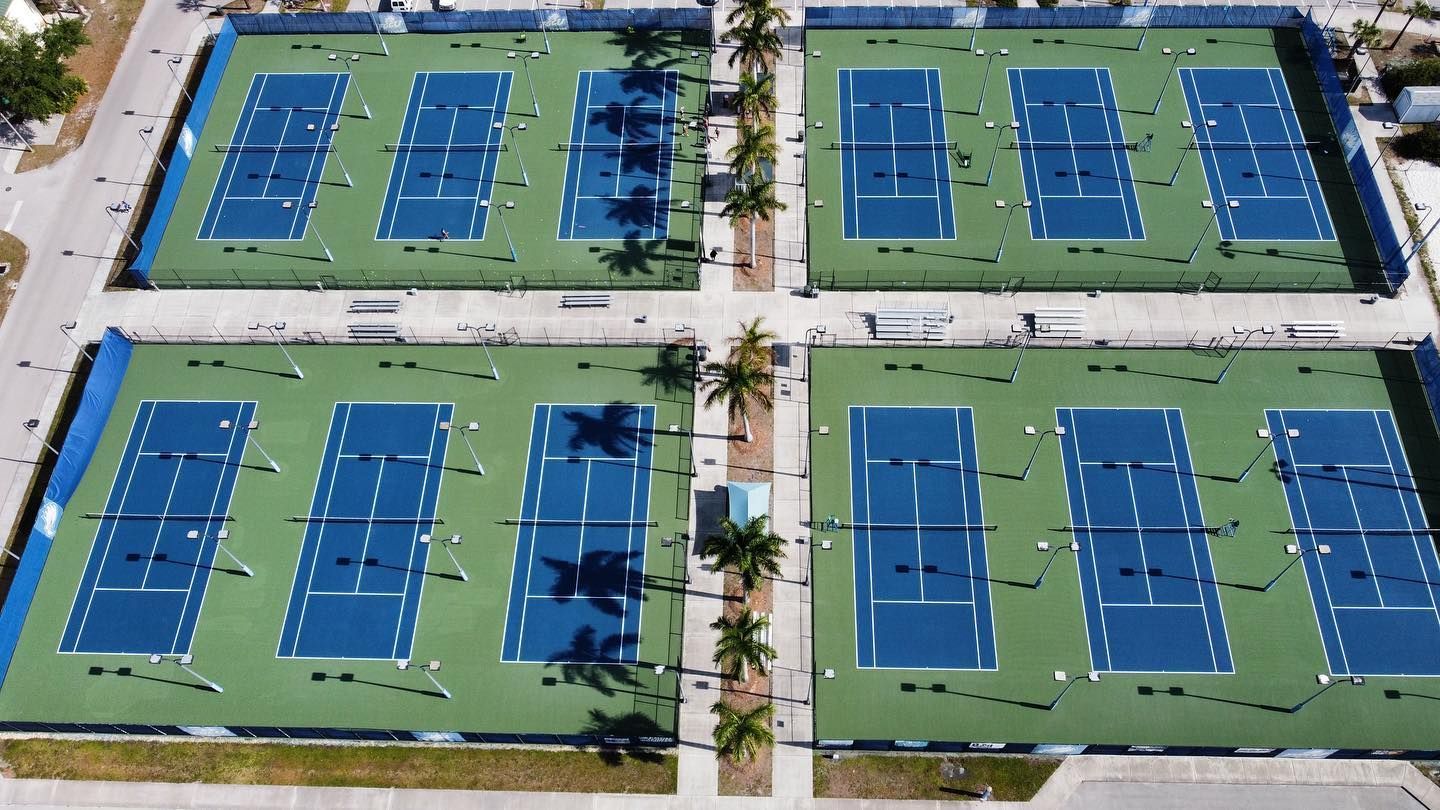 Aerial view of four blue tennis courts with green out-of-bounds areas and palm trees between them.