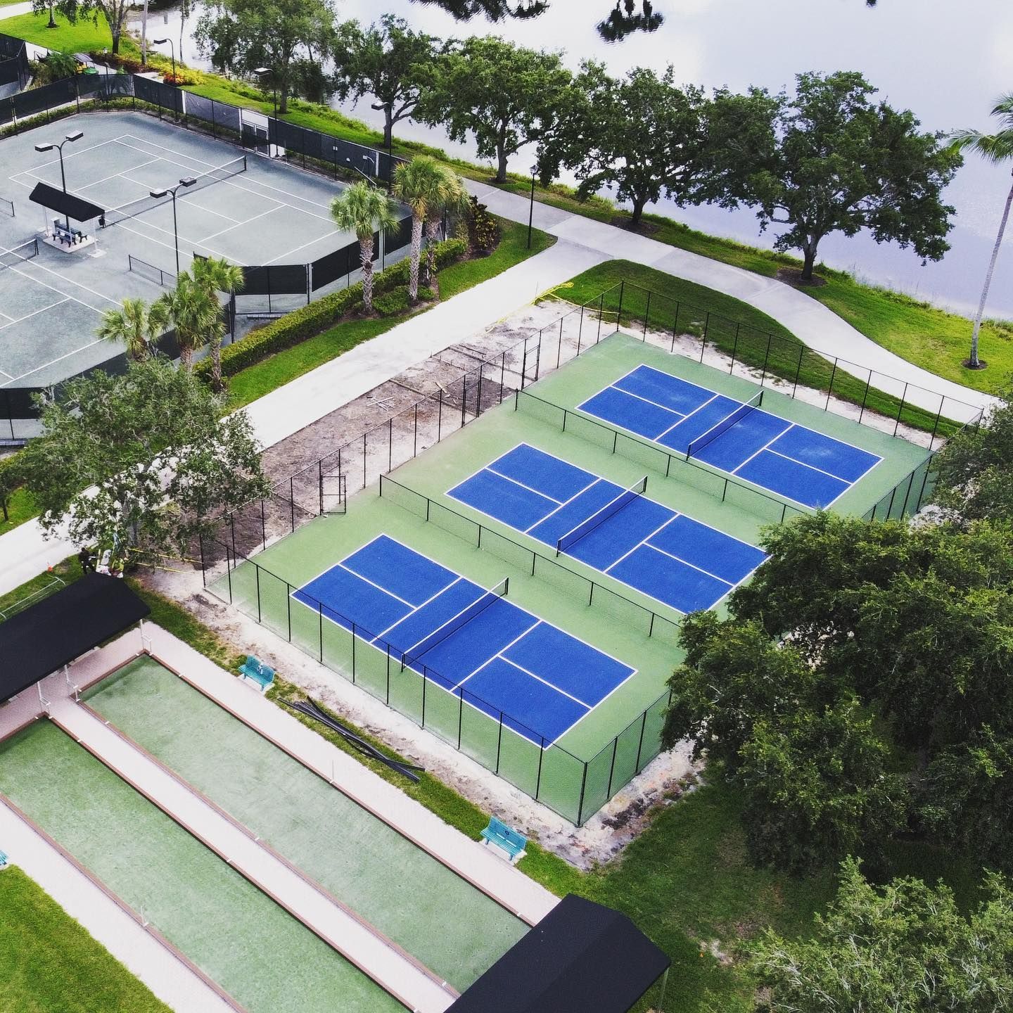 Aerial view of outdoor sports courts with blue and green surfaces beside a tree-lined path and water.