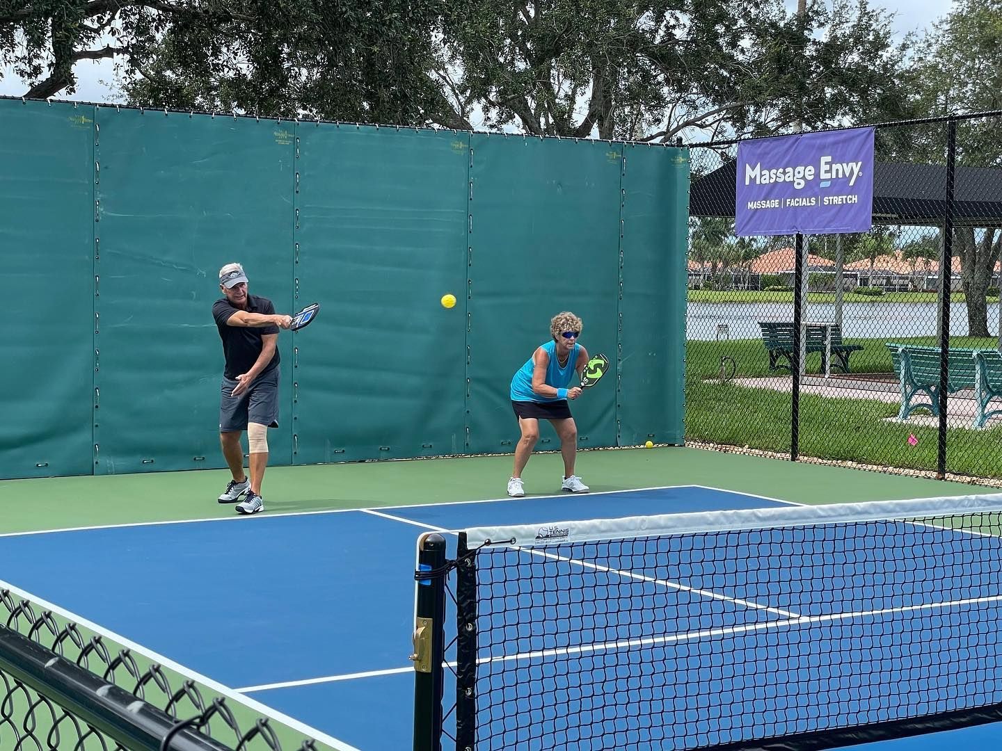 Two players on an outdoor pickleball court, one hitting a yellow ball near a net and green fence.