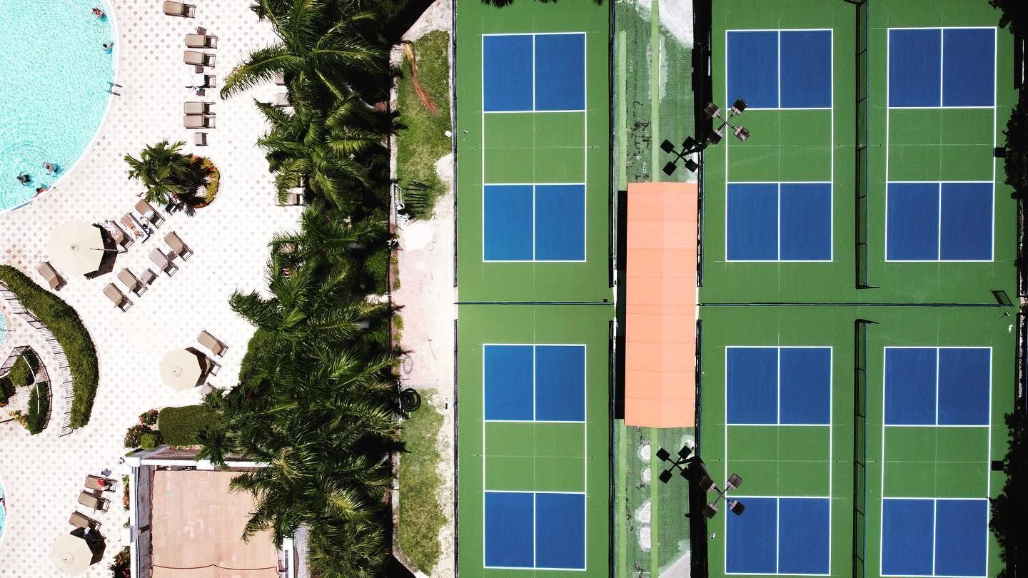 Aerial view of a resort with pools, lounge chairs, palm trees, and adjacent blue tennis courts.