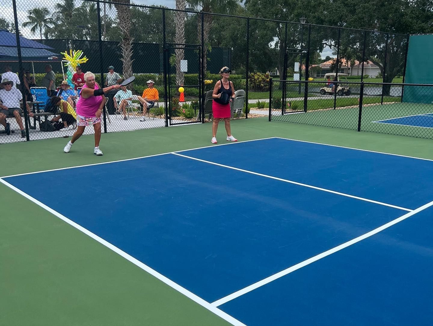 Kids playing pickleball on an outdoor blue court with a black fence and trees in the background