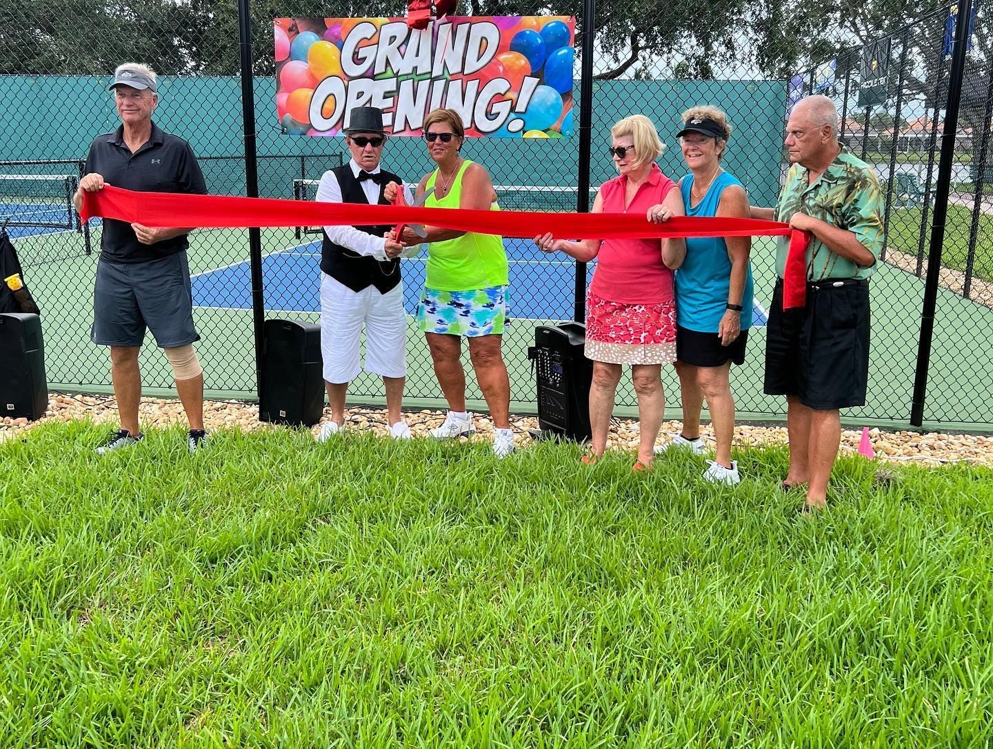 Six people cut a red ribbon at a grand opening sign in a grassy outdoor area.