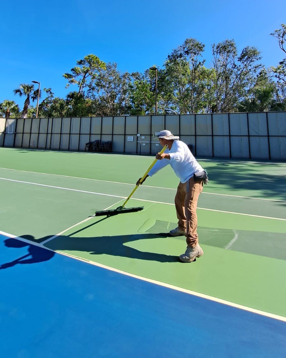 Person painting a tennis court with a roller on a sunny day, using yellow paint on green and blue surface