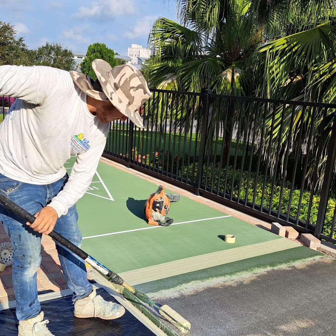 Man trimming a golf green edge beside a small dog sitting on the putting surface, outdoors in sunlight