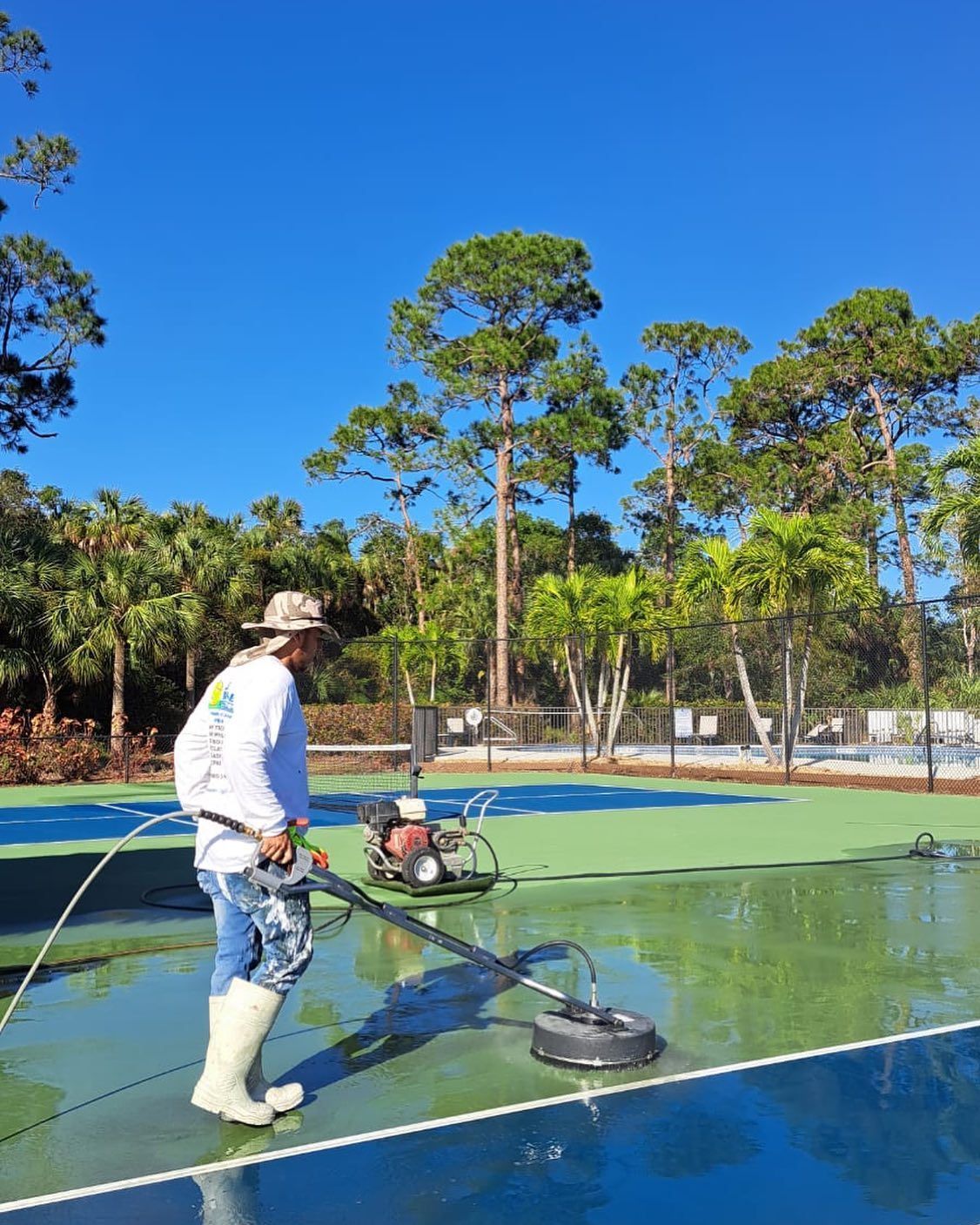 Worker painting a blue court surface with a spray machine under a bright blue sky.