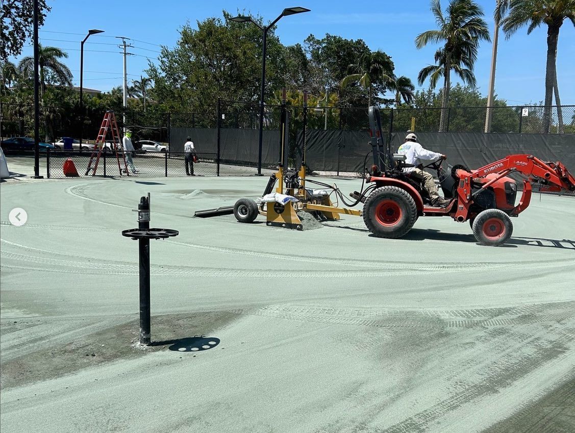 Construction workers operating a red tractor on a newly paved court under palm trees