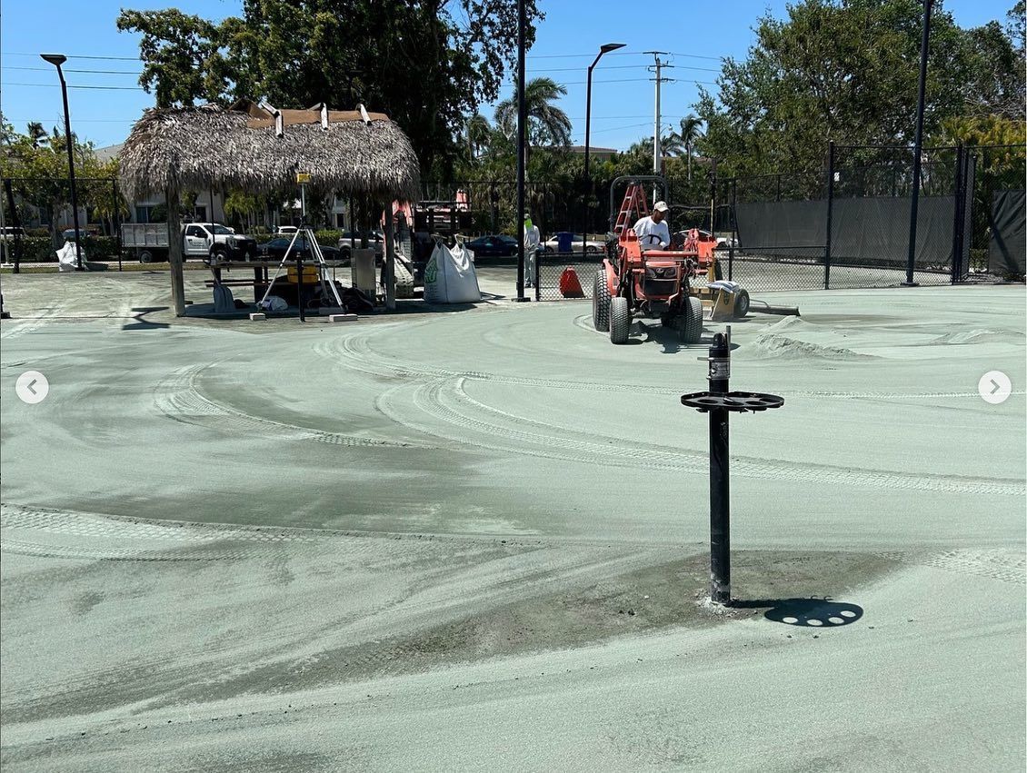 Two red tractors smoothing wet concrete at a fenced outdoor splash pad, with a thatched shade structure nearby