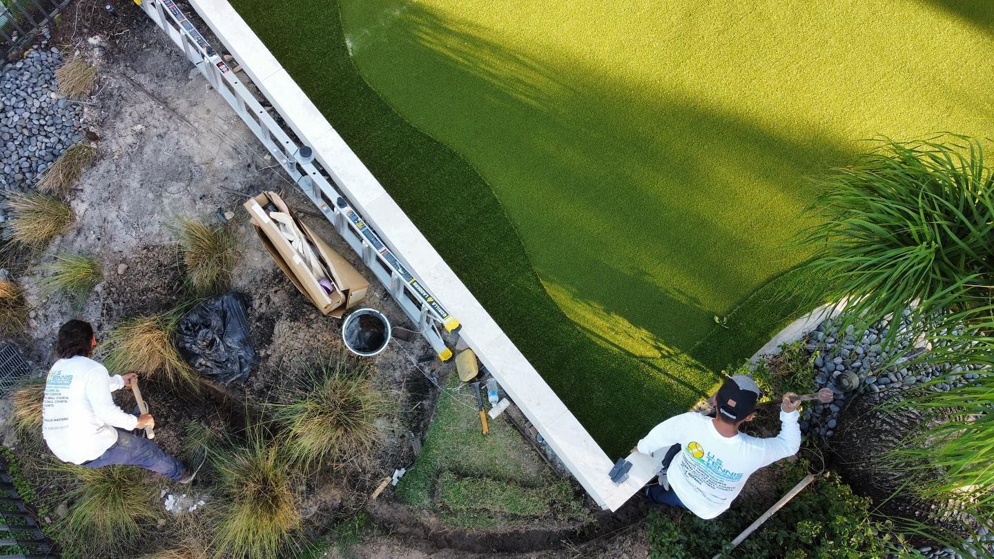 Two workers cleaning a pond edge, one on the bank and one on the curb near green water.