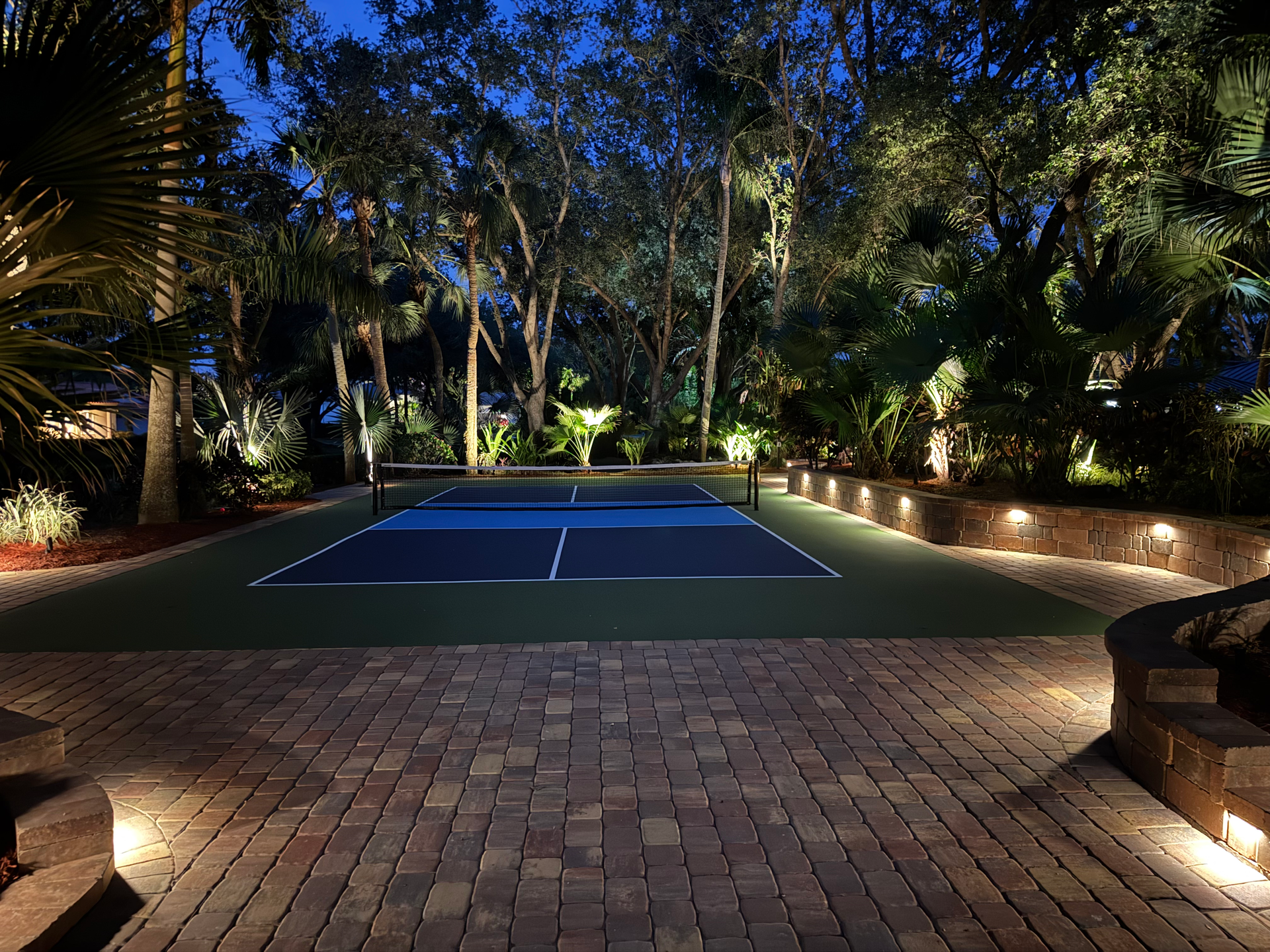Lit tennis court at night, surrounded by trees and pathway lights in a landscaped garden