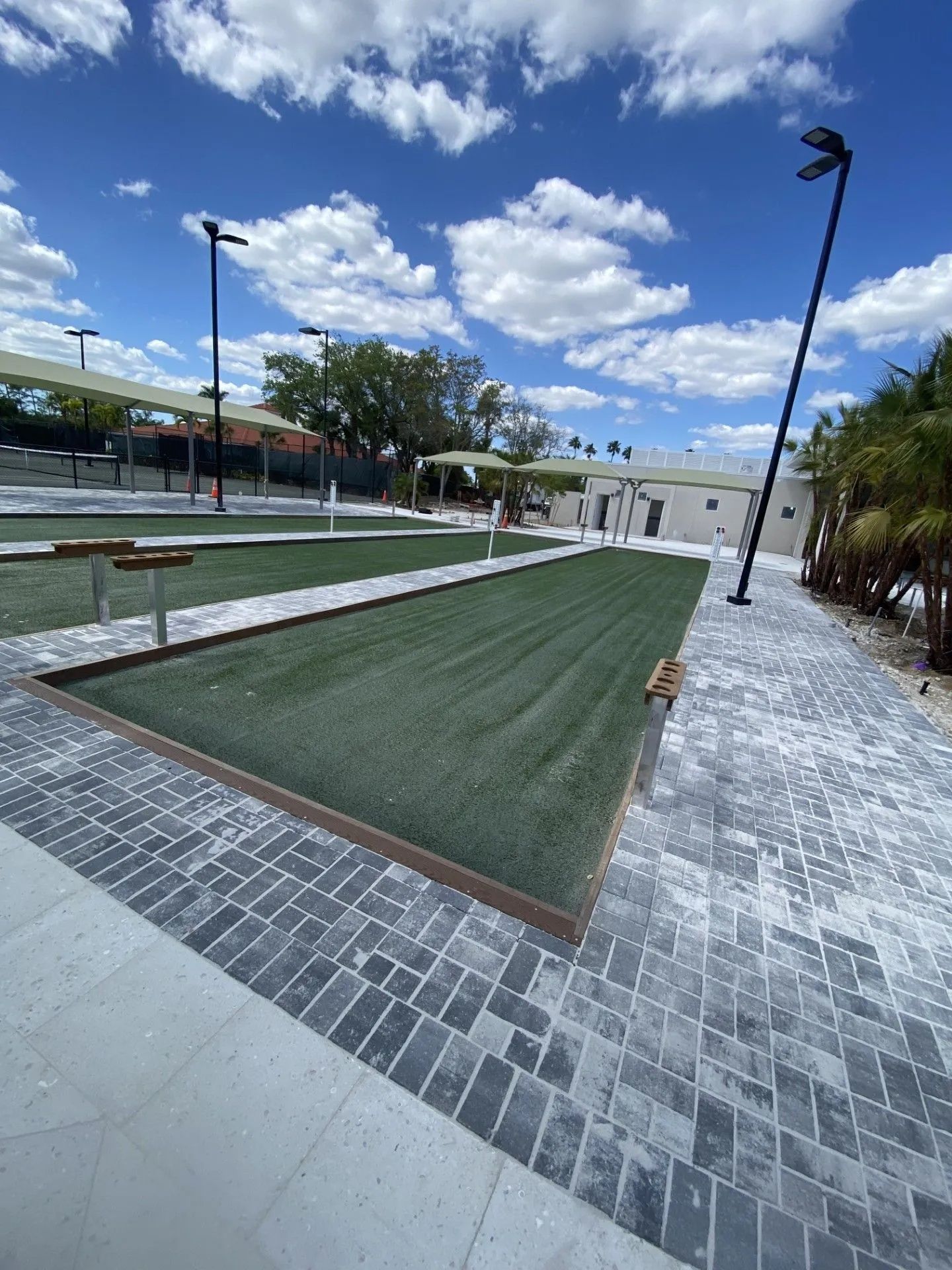 Outdoor park with a long green lawn strip, brick paths, benches, and tall lampposts under a blue sky with clouds