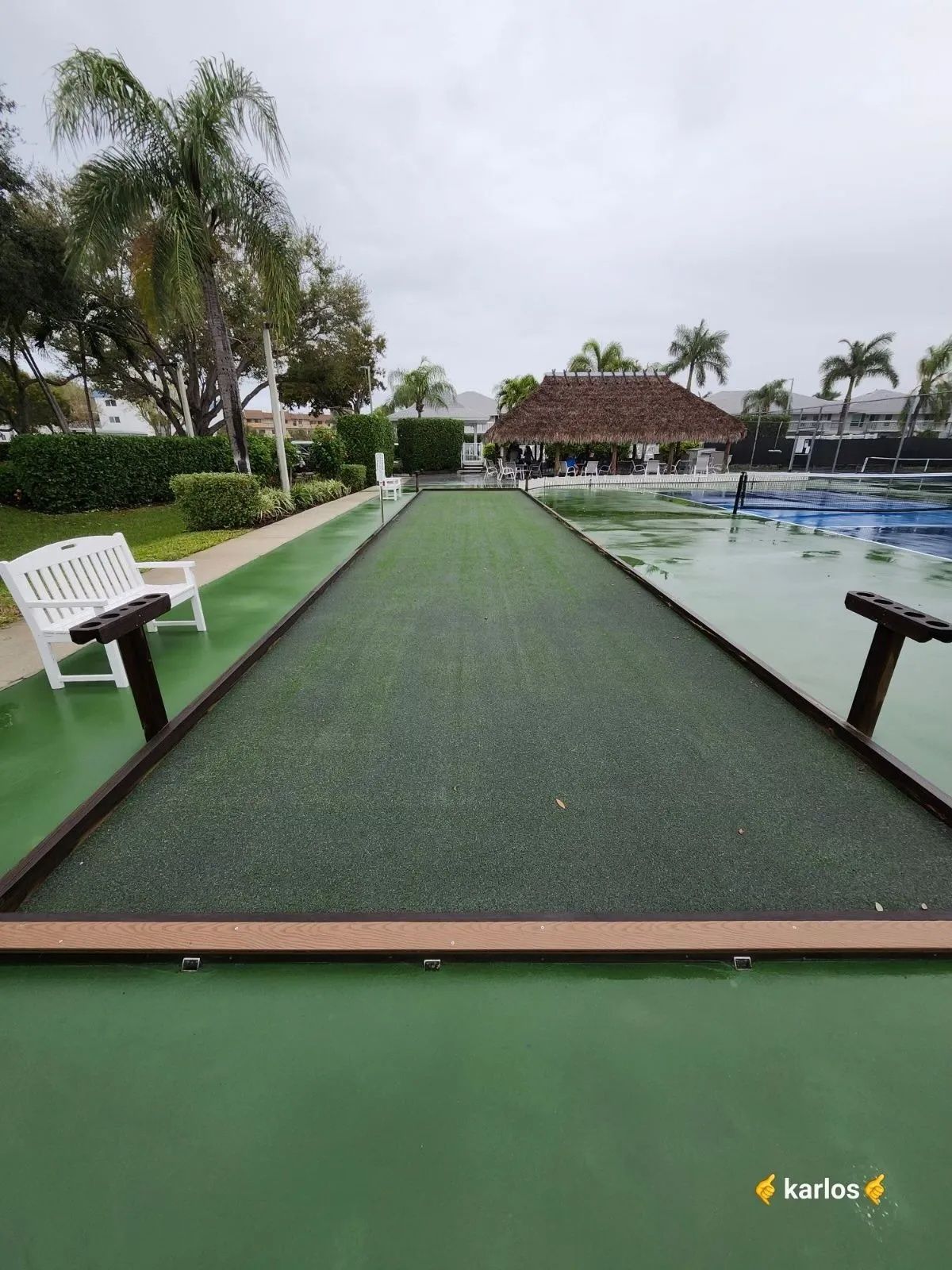 Poolside putting green beside a swimming pool, with palm trees and a thatched gazebo under cloudy skies
