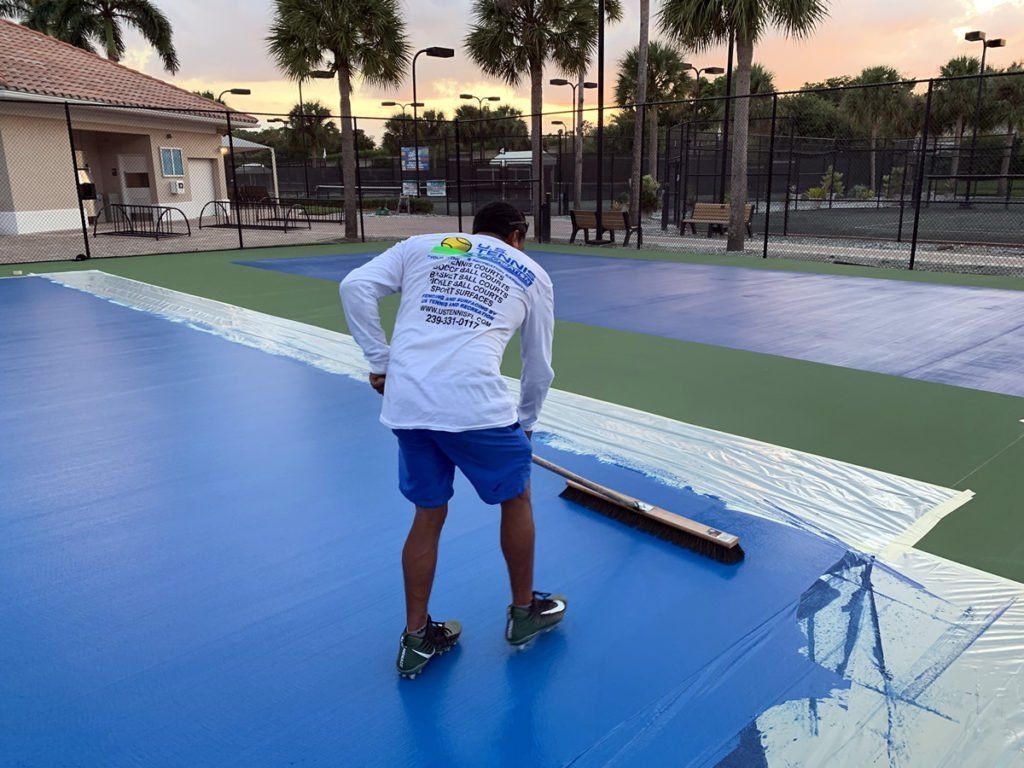 Person on a blue court painting a white line with a roller at dusk.