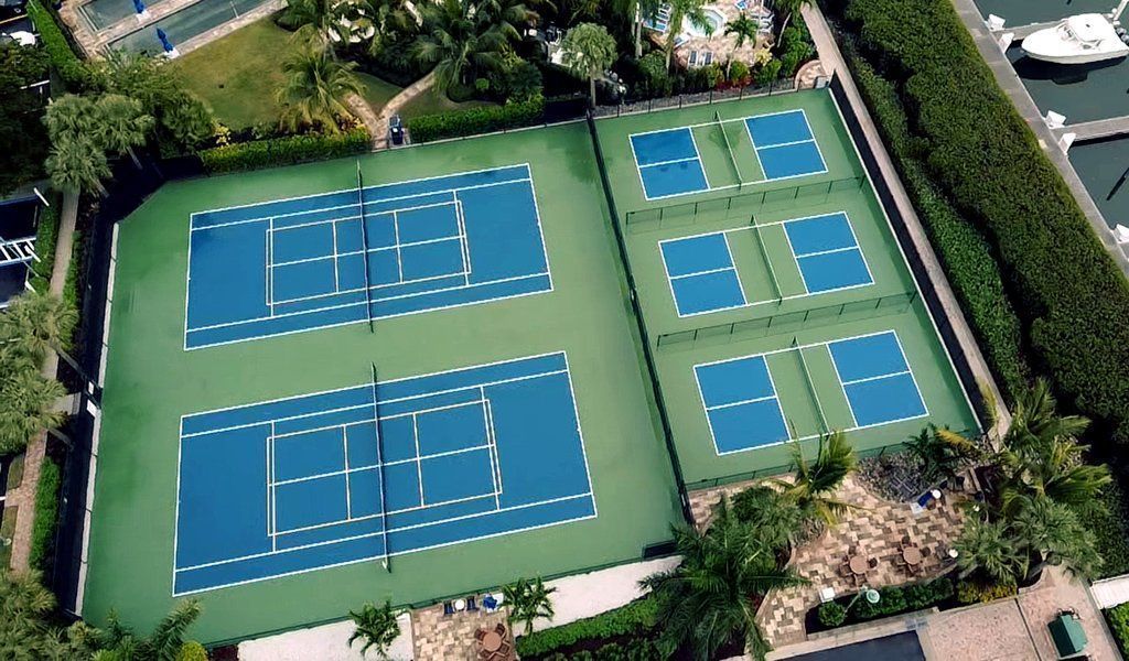 Aerial view of six blue tennis courts with white lines, surrounded by green hedges and trees