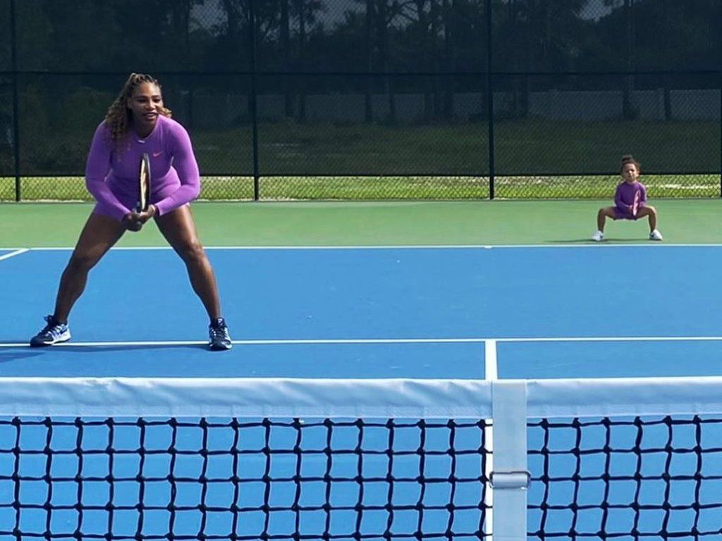 Two tennis players on a blue court, one near the net in a purple top, one seated in the background.