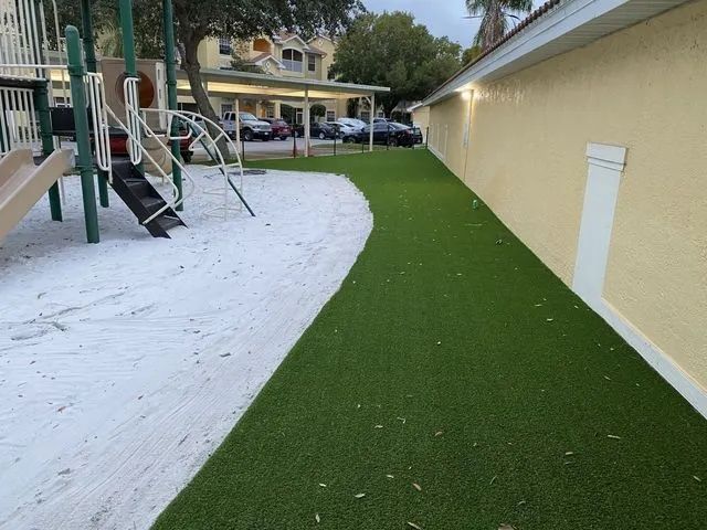 Playground with white ground cover and a green artificial turf path beside a yellow building