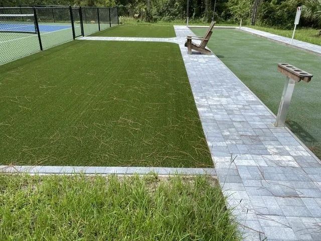 Sports court with artificial turf, a paved walkway, benches, and a fenced tennis court in the background