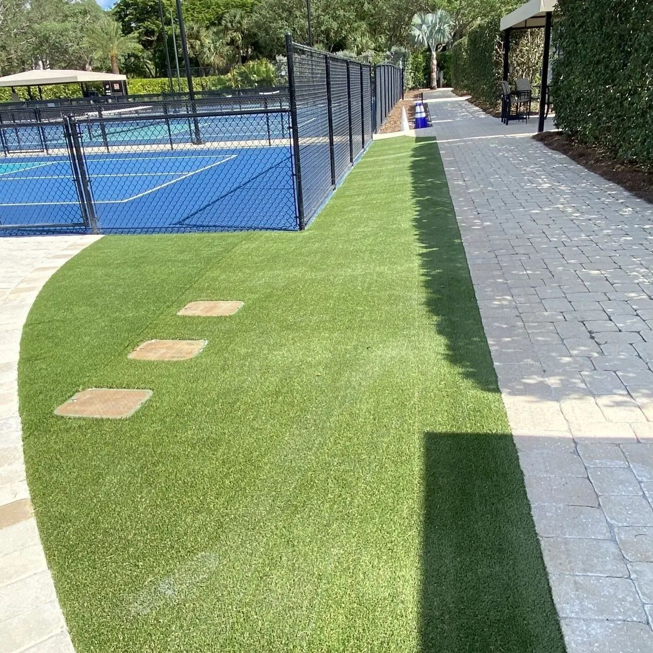 Outdoor poolside walkway beside blue tennis courts, with artificial grass and stone path in bright sun.