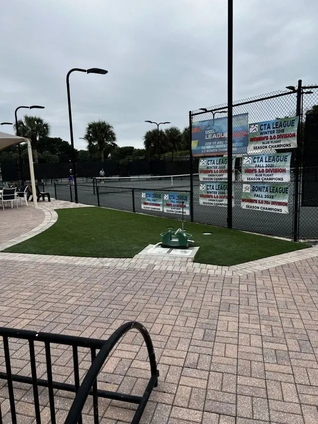 Brick walkway beside a fenced sports court with banners, lamp posts, and a small grassy island