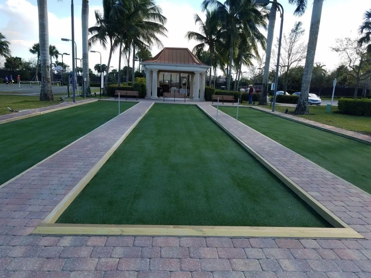 Palm-lined walkway leading to a small pavilion in a landscaped park, with manicured grass and stone paths.
