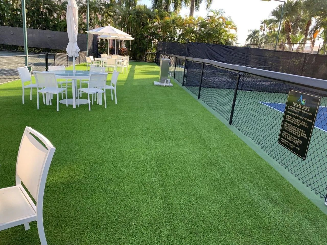 Outdoor lawn with white patio tables and chairs, a black chain-link fence, and umbrellas near a pool area