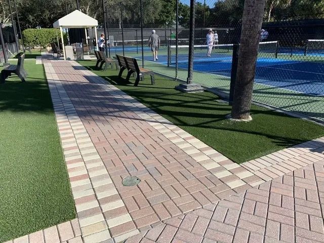 Brick walkway beside a tennis court, with benches, palm trees, and a white gazebo in a sunny park.