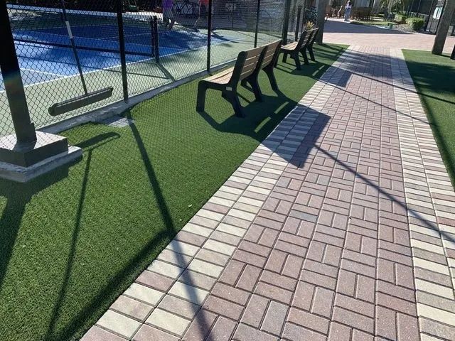 Outdoor walkway beside a fenced pool with black benches, artificial turf, and long shadows on brick pavers