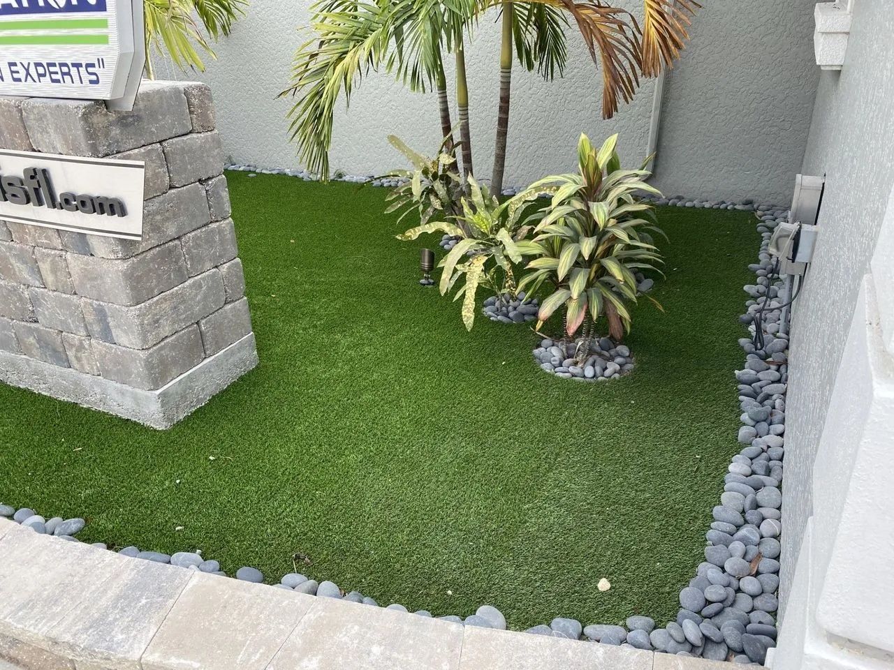Small landscaped corner with artificial grass, potted plants, stone edging, and a sign on a wall