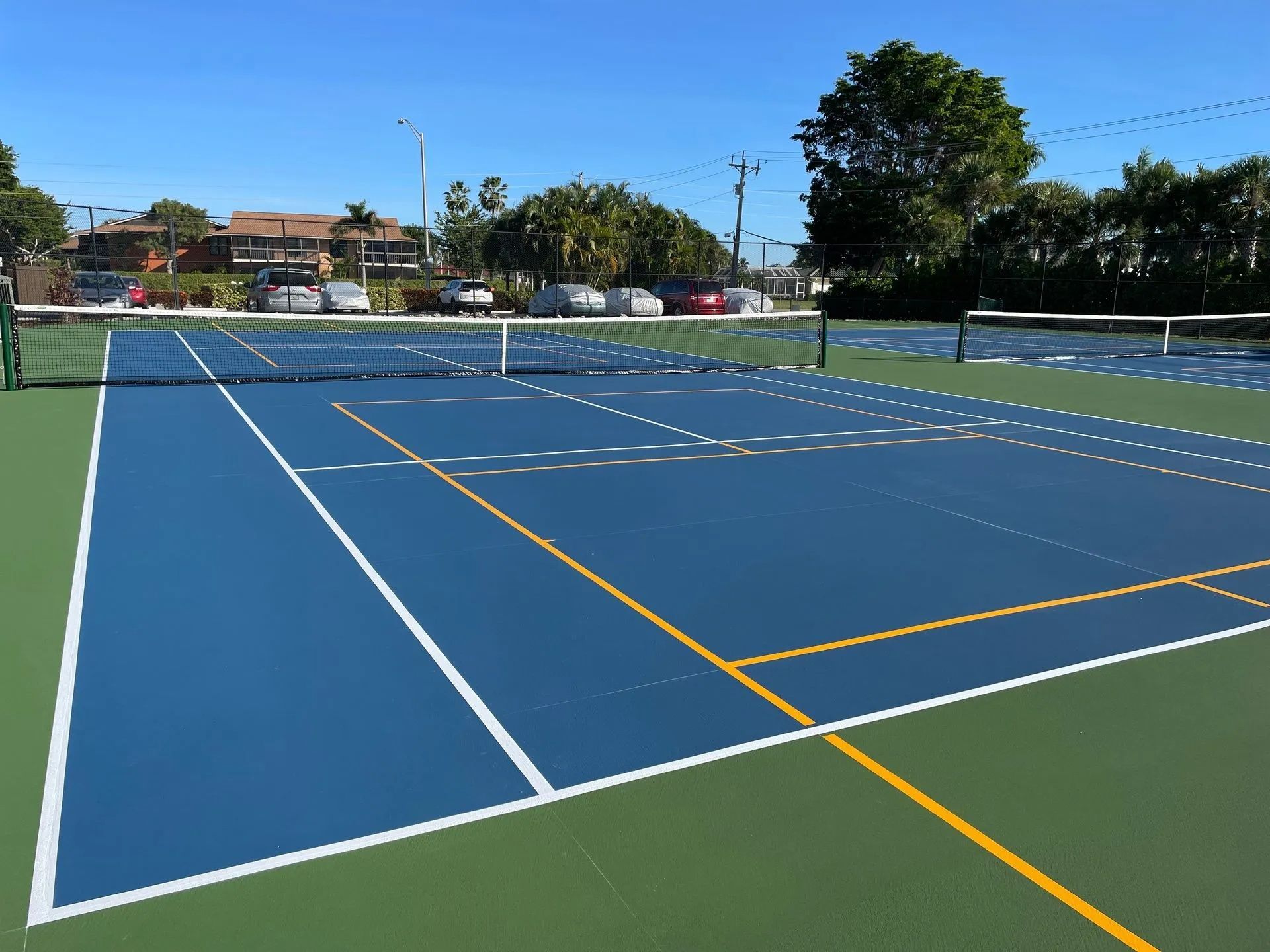 Blue outdoor tennis court with white and yellow lines under a clear sky, surrounded by trees and houses