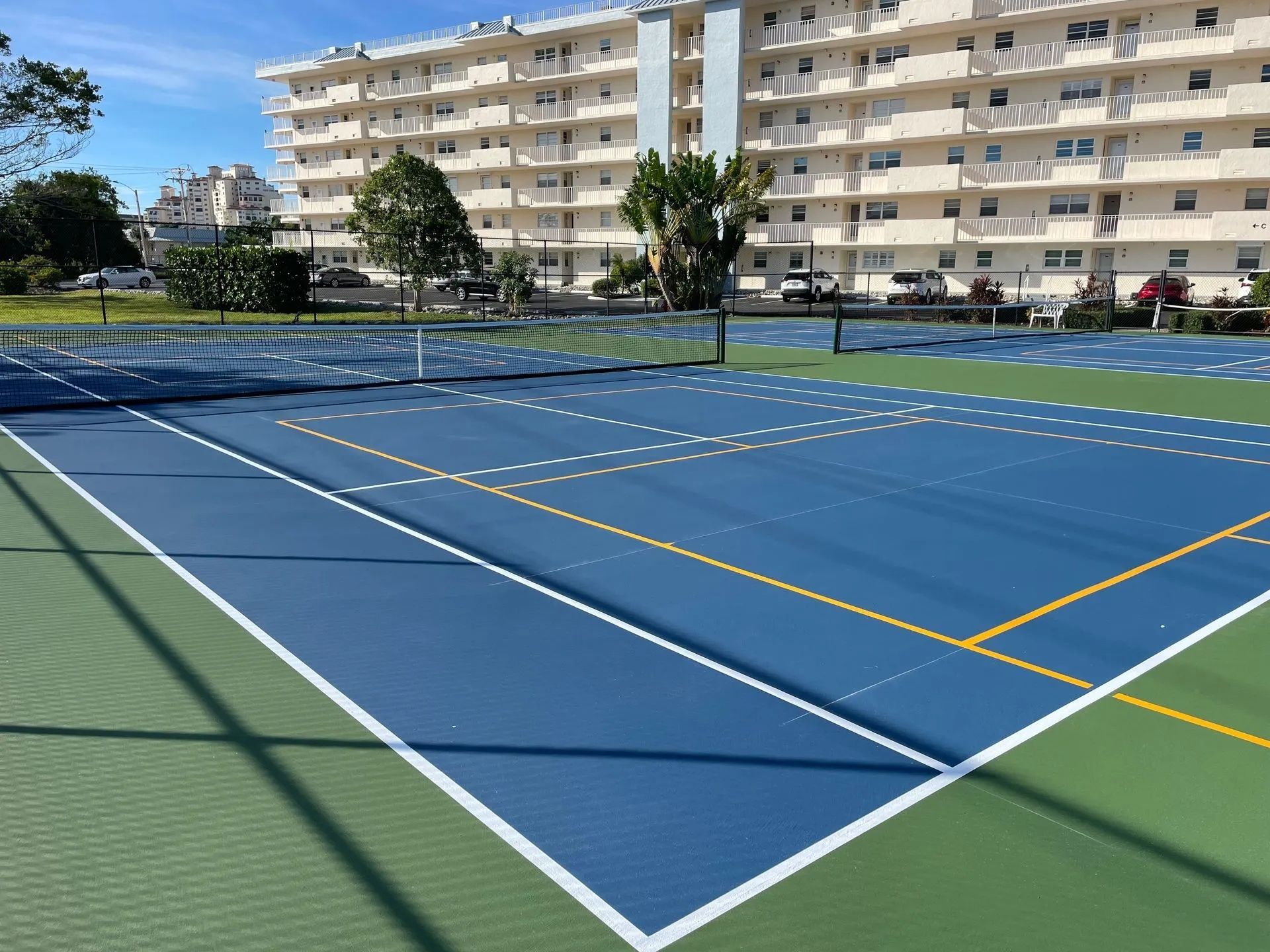 Blue tennis court with green outer area, surrounded by a fence and apartment buildings in the background